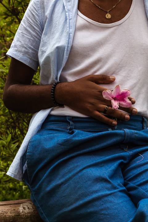 Hugo em frente a parede vermelha de tijolos no parque do carmo, na capital de são paulo. Em frente aos arbustos, com uma flor em frente a sua barriga, enquadramento parcial do corpo.'