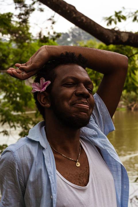 Hugo no parque do carmo, na capital de são paulo. Com uma flor na cabeça, enquanto sorri leve, enquadramento mais em seu rosto, em frente ao lago.'
