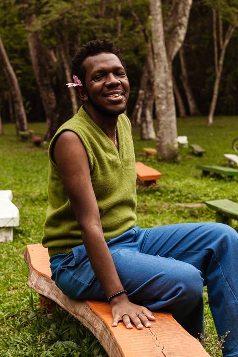 Hugo no parque do carmo, na capital de são paulo. Com uma flor na cabeça, enquanto sorri leve, sentado nos bancos coloridos.'