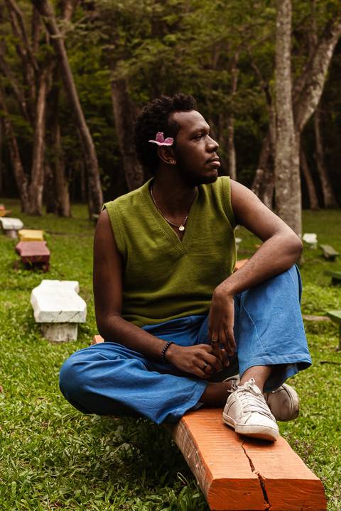 Hugo no parque do carmo, na capital de são paulo. Com uma flor na cabeça, enquanto sorri leve, sentado nos bancos coloridos, olhar ao horizonte, uma das pernas em cima do banco.'