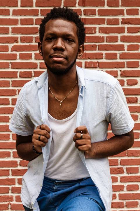 Hugo em frente a parede vermelha de tijolos no parque do carmo, na capital de são paulo. Segurando jaqueta, olhando diretamente para mim.'