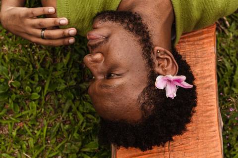 Hugo no parque do carmo, na capital de são paulo. Com uma flor na cabeça, enquanto olha para a lateral com a mão sobre o ombro, deitado nos bancos coloridos.'