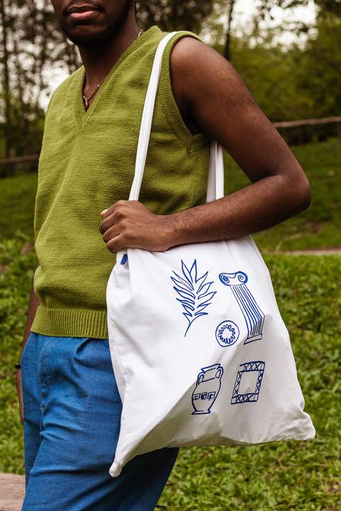 Hugo no parque do carmo, na capital de são paulo. Com uma flor na cabeça, enquanto sorri leve, em pé ao lado dos bancos coloridos, pegando parcial do seu corpo.'