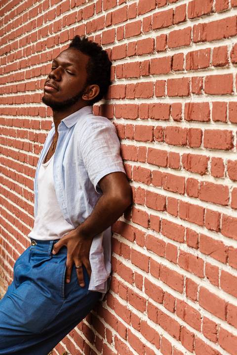 Hugo em frente a parede vermelha de tijolos no parque do carmo, na capital de são paulo. Com as mãos sobre a calça, encostado na parede de tijolos vermelhos, olhos fechados.'