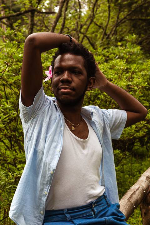 Hugo em frente a parede vermelha de tijolos no parque do carmo, na capital de são paulo. Em frente aos arbustos, com uma flor na cabeça.'