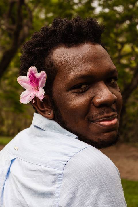 Hugo em frente a parede vermelha de tijolos no parque do carmo, na capital de são paulo. Em frente aos arbustos, com uma flor na cabeça, enquanto sorri leve, enquadramento mais em seu rosto.'