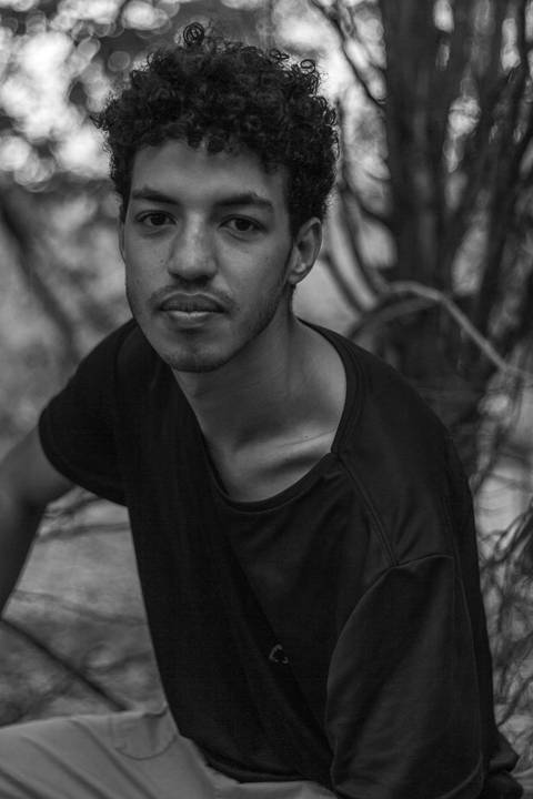 Isaque em meio as árvores, época de outono no parque do carmo, folhas ao seu redor, enquadramento mais fechado, apoiado com os joelhos no chão.'