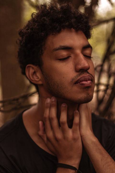 Isaque em meio as árvores, época de outono no parque do carmo, folhas ao seu redor, enquadramento mais fechado, enquanto coloca as mãos ao redor do pescoço. '