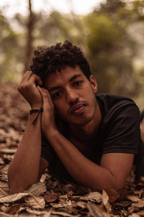 Isaque em meio as árvores, época de outono no parque do carmo, folhas ao seu redor, enquadramento mais fechado, deitado no chão, apoiando a cabeça sobre os braços, sério, olhando direto para mim.'