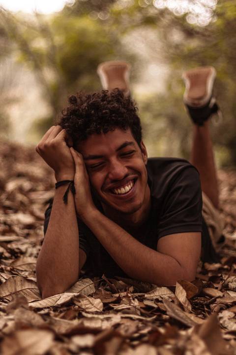 Isaque em meio as árvores, época de outono no parque do carmo, folhas ao seu redor, enquadramento mais fechado, deitado no chão, apoiando a cabeça sobre os braços, sorrindo espontâneo, olhando direto para mim.'