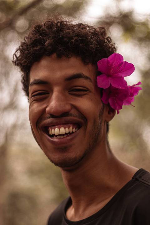 Isaque em meio as árvores, época de outono no parque do carmo, folhas ao seu redor, enquadramento mais fechado, sentado no chão, sorrindo espontâneo, olhando direto para mim, ainda mais próximo, enquadramento fechado no rosto.'