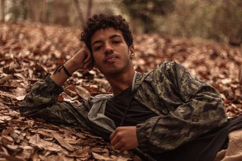 Isaque em meio as árvores, época de outono no parque do carmo, folhas ao seu redor, enquadramento mais fechado, sentado no chão, sorrindo espontâneo, olhando direto para mim, deitado no chão.'