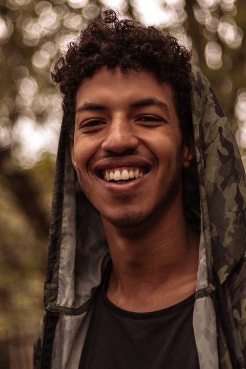 Isaque em meio as árvores, época de outono no parque do carmo, folhas ao seu redor, enquadramento mais fechado, sentado no chão, sorrindo espontâneo, olhando direto para mim, capuz da blusa.'