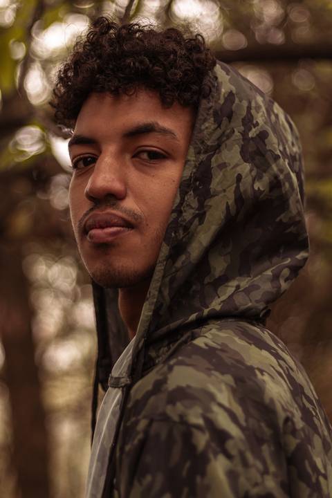 Isaque em meio as árvores, época de outono no parque do carmo, folhas ao seu redor, enquadramento mais fechado, sentado no chão, olhando direto para mim, capuz da blusa, sério.'