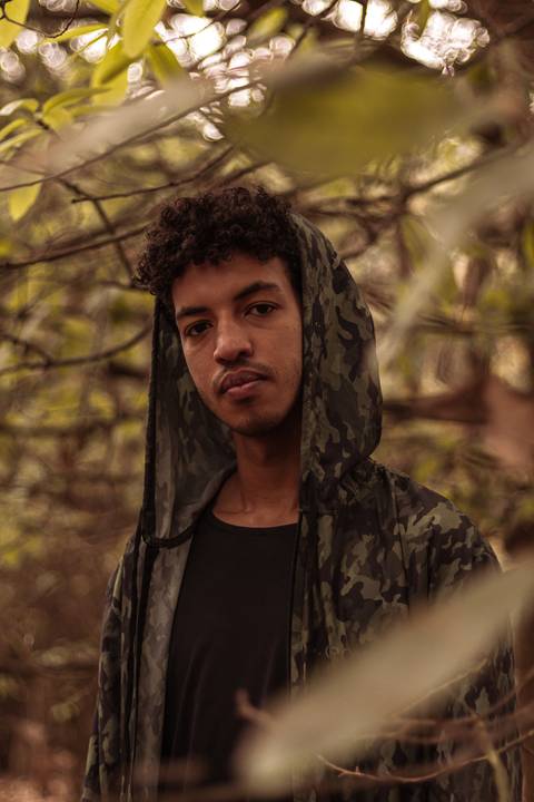 Isaque em meio as árvores, época de outono no parque do carmo, folhas ao seu redor, enquadramento mais fechado, sentado no chão, olhando direto para mim, capuz da blusa, sério e de frente para a câmera.'