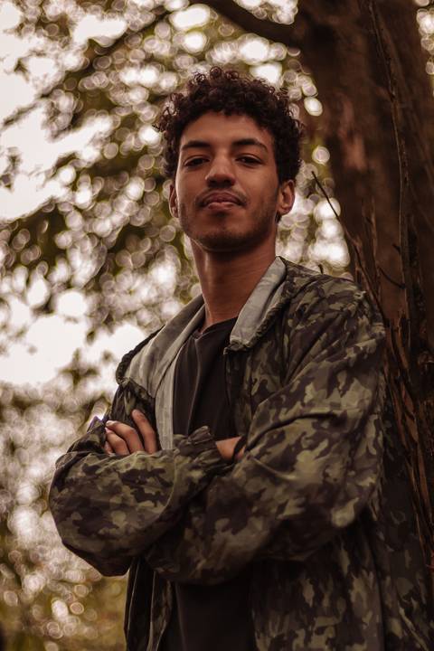 Isaque em meio as árvores, época de outono no parque do carmo, folhas ao seu redor, enquadramento mais fechado, sentado no chão, olhando direto para mim, encostado na árvore, com os braços cruzados em frente ao peito.'