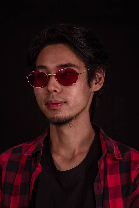 Jorge em estúdio na capital de são paulo, sentado em frente ao fundo preto, camiseta xadez, olhando em direção para a câmera, enquadramento mais próximo, óculos escuros, rosto virado a 45 graus, sorrido leve.'