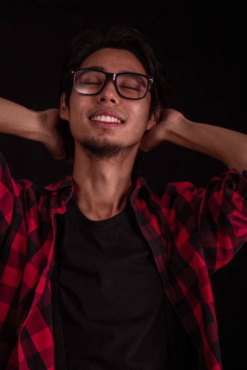 Jorge em estúdio na capital de são paulo, sentado em frente ao fundo preto, camiseta xadez, olhando em direção para a câmera, enquadramento mais próximo, óculos escuros, sorrindo com as mãos ao redor da cabeça.'