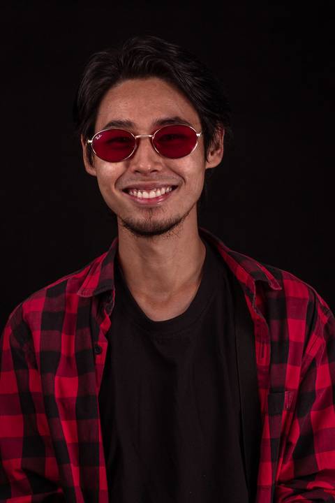 Jorge em estúdio na capital de são paulo, sentado em frente ao fundo preto, camiseta xadez, olhando em direção para a câmera, enquadramento mais próximo, óculos escuros, enquanto sorri espontâneo.'