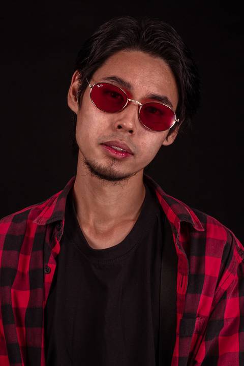 Jorge em estúdio na capital de são paulo, sentado em frente ao fundo preto, camiseta xadez, olhando em direção para a câmera, enquadramento mais próximo, óculos escuros, rosto inclinado, mais sensual.'