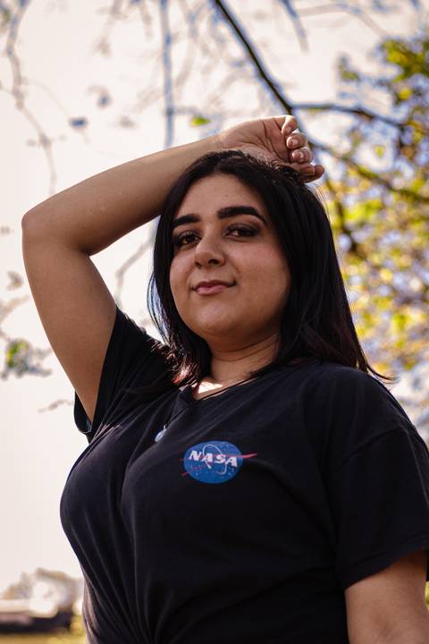 Kellen, no parque do Ibirapuera, com árvores e folhagens ao fundo, com a mão sobre o cabelo, olhando diretamente para a câmera.'