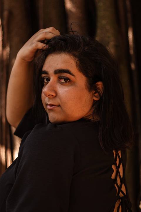 Kellen a frente de uma árvore no parque do Ibirapuera, olhando séria diretamente para a câmera, com vestido preto e com as mãos sob os cabelos negros.'