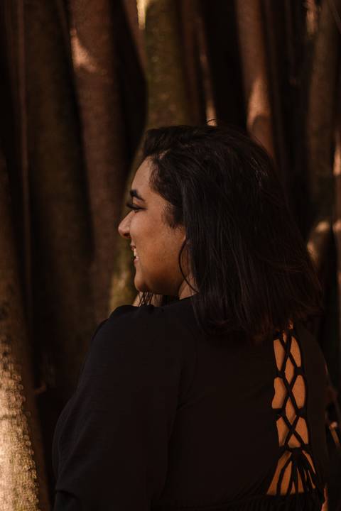 Kellen olhando além do horizonte, sorrindo, quase de costas para a câmera, em frente a uma árvore no parque do Ibirapuera.'