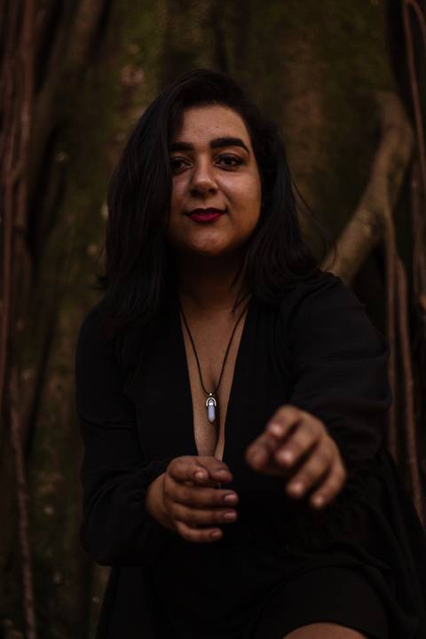 Kellen estendendo os braços em direção a câmera fotográfica, olhando diretamente para ela, com fundo de árvores no parque do Ibirapuera.'