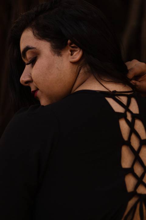 Kellen de costas, mostrando detalhes do seu vestido preto, com uma das mãos em seus cabelos negros, com a lateral do seu rosto em foco.'