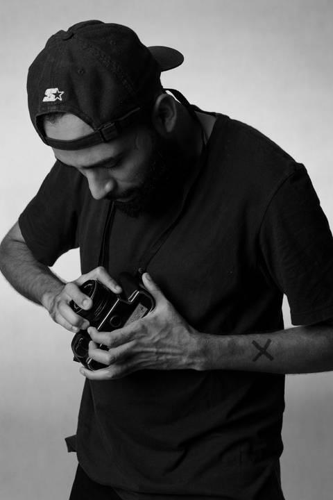 Kevin, em estúdio com fundo branco, olhando diretamente para a câmera fotográfica em suas mãos, manuseando a mesma, na intenção de girar a lente da câmera.'