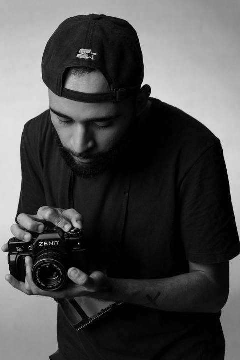 Kevin, em estúdio com fundo branco, olhando diretamente para a câmera fotográfica em suas mãos, analisando minuciosamente cada detalhe dela.'