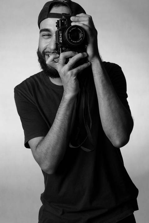 Kevin, em estúdio com fundo branco, olhando através da câmera em suas mãos, como se fosse fotografar a fotógrafa, sorrindo por trás do seu equipamento.'