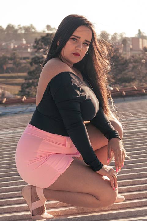 Lethícia ajoelhada olhando diretamente para a câmera, sob telhado da sua casa, com fundo de casas e céu.'