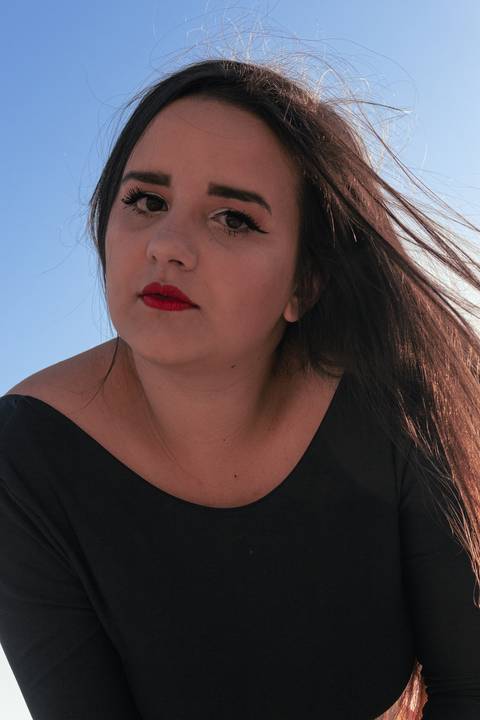 Fundo totalmente de céu azul, rosto inclinado para a lateral diretia, olhar fixo na direção da câmera, Lethícia sendo fotografada de baixo, cabelos soltos ao vento.'