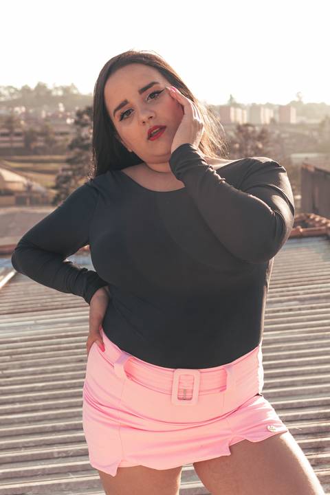 Lethícia com uma mão na cintura e outra em seu rosto, no telhado da sua casa, com fundo de casas e céu azul.'