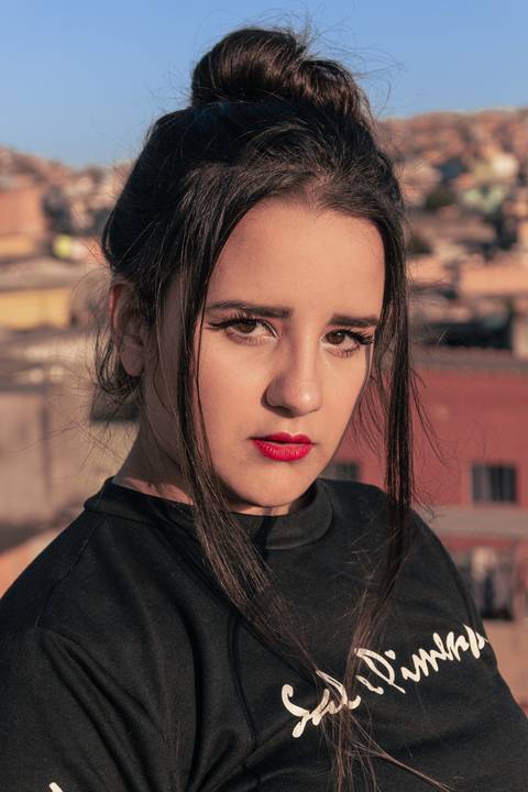 Lethícia com os cabelos amarrados em coque, com mexas soltas a frente do rosto, olhando diretamente para a câmera, séria.'
