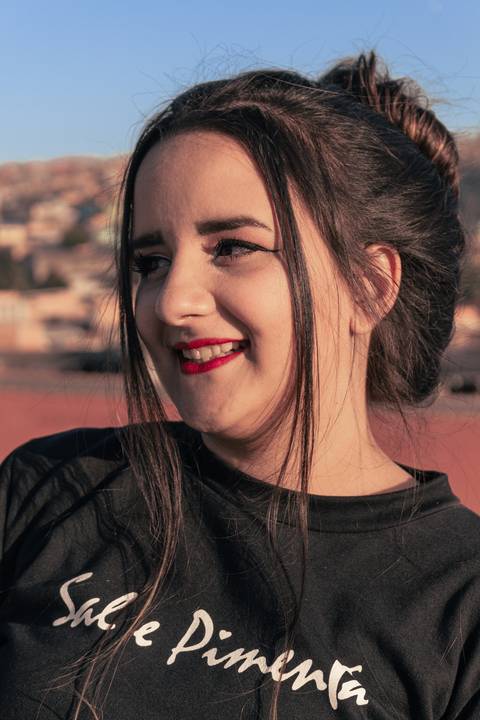 Lethícia com os cabelos amarrados em coque, com mexas soltas a frente do rosto, olhando para além do horizonte, sorrindo.'