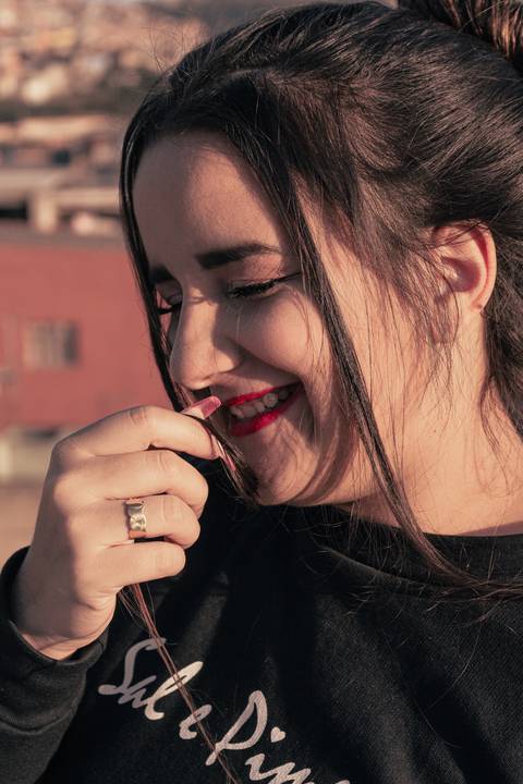 Lethícia com os cabelos amarrados em coque, com mexas soltas a frente do rosto, os os olhos fechados, sorrindo, com a mão próxima ao rosto.'