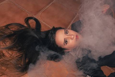 Lethícia fumando narguilé dentro de sua casa, deitada no chão, com fumaça ao seu redor, cabelos lisos longos espalhados pelo chão. olhar diretamente para a câmera.'