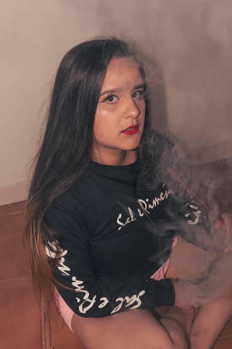 Lethícia sentada no chão da sala de sua casa fumando narguilé, olhando diretamente para a câmera, com fumaça ao seu redor.'
