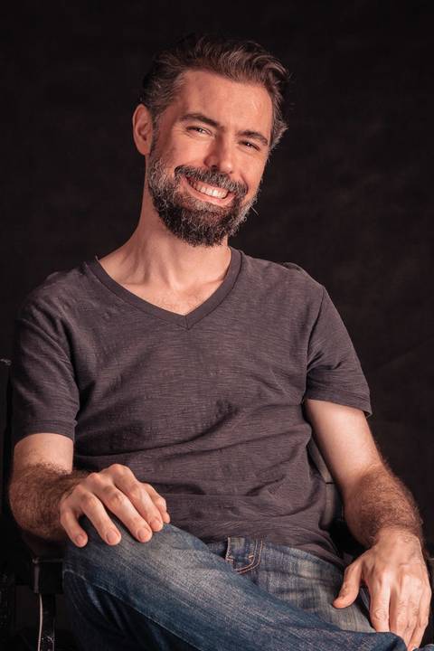 Luís sentado em cadeira dentro de estúdio com fundo preto, sorrindo para a câmera.'