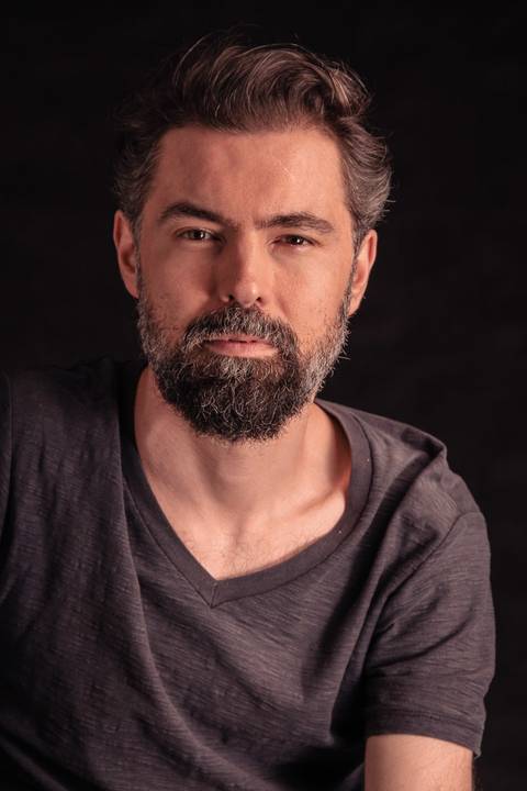 Luís, olhando diretamente para a câmera, sério, sentado na cadeira em estúdio, com fundo preto.'
