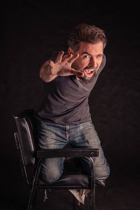 Luís em cima da cadeira em estúdio com fundo preto, fazendo uma pose como se quisesse alcançar algo.'