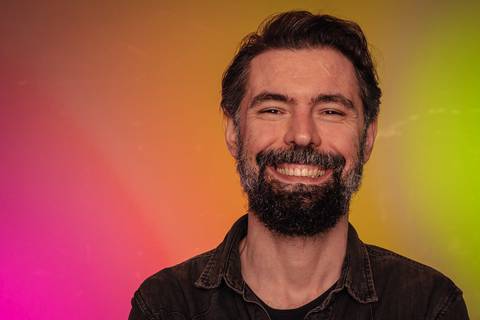 Luís sorrindo em estúdio com fundo de parede colorida.'