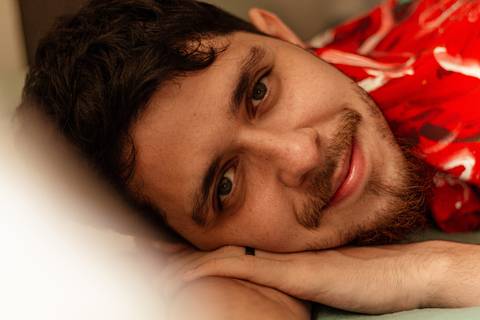 Matheus, com o olhar direcionado para a câmera, com sorrido leve, deitado em sua cama, com cortina pegando uma parte da imagem.'