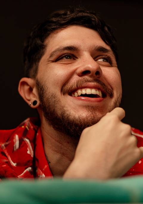 Matheus olhando além da câmera sorrindo espontâneo, deixado de bruços sobre a sua cama.'
