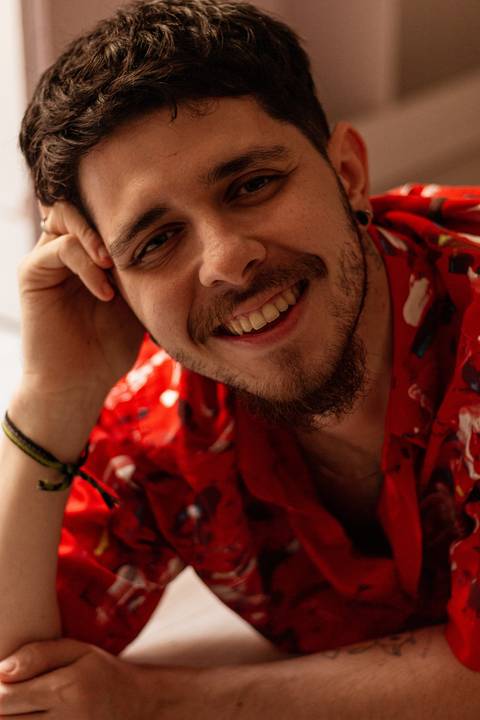 Matheus, deitado de bruços no chão de sua casa, apoiando a cabeça em um dos braços, sorrindo espontâneo para a câmera.'