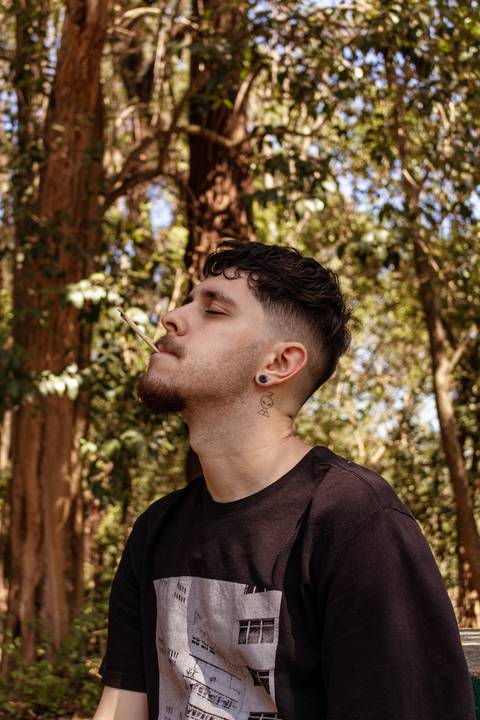 Matheus com árvores ao seu redor no parque do Ibirapuera, na capital de são paulo, fumando cigarro de palha, olhos fechados, cabeça para o alto.'