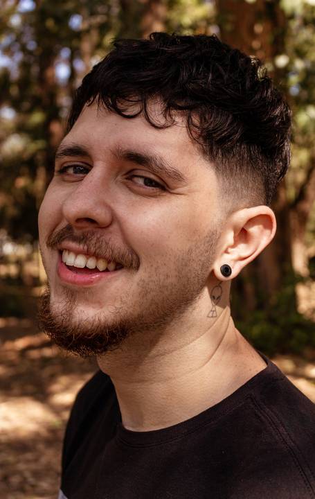 Matheus com árvores ao seu redor no parque do Ibirapuera, na capital de são paulo, enquadramento fechado em seu rosto, sorrindo para a câmera.'