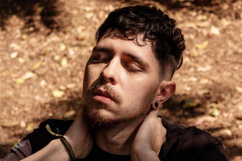 Matheus com árvores ao seu redor no parque do Ibirapuera, na capital de são paulo, enquadramento fechado em seu rosto, olhos fechados, cabeça para o céu, mãos no pescoço.'